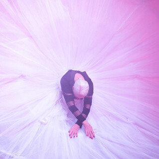 A person in a long, fluffy coat poses on a red carpet. They hold black balloons in their hand while smoke and lights create a dramatic atmosphere. | © Alexi Pelekanos