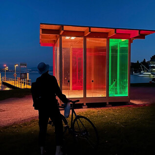 A modern, illuminated glass cabin stands on the shore of a body of water. A person with a bicycle observes the colored lights at night. | © DdePerrot