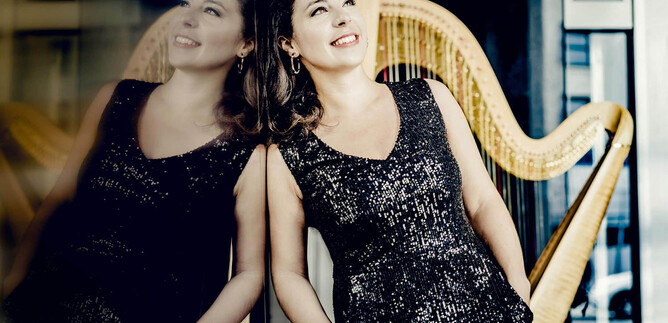 An elegant woman in a glittering dress smiles against a wall. In the background, a harp is visible. | © Andrej Grilc