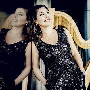 An elegant woman in a glittering dress smiles against a wall. In the background, a harp is visible. | © Andrej Grilc