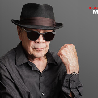An older man is wearing a black hat and sunglasses. He poses confidently in front of a gray background. | © Alejandro Saldana