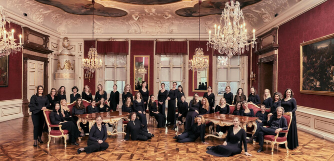 A group of women in elegant clothing sits and stands in a magnificent room with chandeliers. The room is richly decorated with red walls and a detailed ceiling fresco. | © Bachchor Salzburg_Foto: Andreas Hechenberger