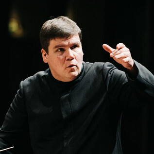 A conductor stands in the center and leads the orchestra. He makes clear gestures with his hand. | © Roberts Blaubuks