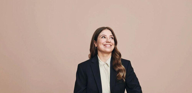 A young woman is sitting smiling in a black suit. The background is in a soft beige tone. | © Veikko Kähkönen