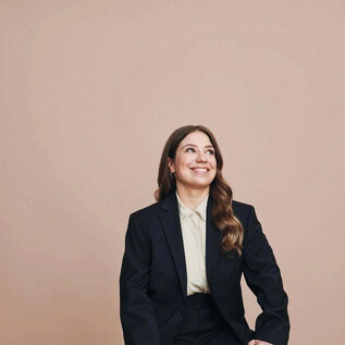A young woman is sitting smiling in a black suit. The background is in a soft beige tone. | © Veikko Kähkönen