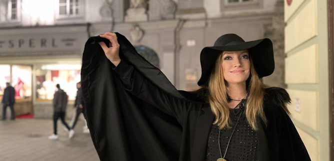 Une femme élégante en manteau noir et grand chapeau se tient dans une rue de la ville la nuit. En arrière-plan, des lumières et des vitrines sont visibles. | © Sabine Rath