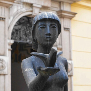 An elegant statue of a woman with short hair, displaying an open palm. In the background, there is a historic building with a decorative door. | © Mag. Christoph Koca