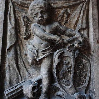 An antique relief of a robe-wearing, winged angel holding a harp. The background features a meticulously designed wall with additional decorative elements. | © Mag. Christoph Koca