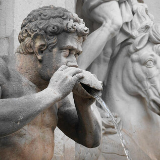 An old statue of a man pouring water from a drinking vessel. In the background, there is a horse with a detailed head. | © Mag. Christoph Koca