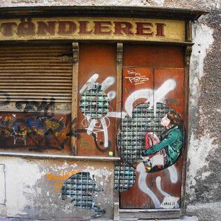 A graffiti-covered shop with the inscription "Töndlerei". A figure sits on the wall, surrounded by colorful artwork. | © Mag. Christoph Koca