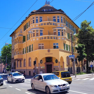 A yellow building with stylish windows and balconies at a street corner. Several cars are passing by, and the surroundings are green and sunny. | © Christoph Koca