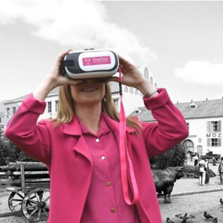 A woman is wearing a virtual reality headset and looking curiously. In the background, historical buildings and people can be seen. | © Sabine Rath