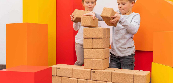 Two children are building a structure with wooden blocks. Colorful geometric shapes are visible in the background. | © Salzburg Museum