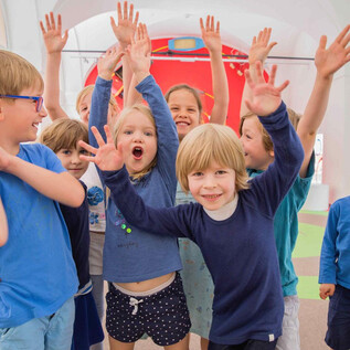 A group of cheerful children stands in a colorful room, raising their hands. They are all wearing blue clothing and radiating joy. | © Eva trifft_Fotografie