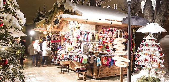 A festive Christmas market with a wooden stall offering colorful handcrafted items. The surroundings are covered in snow and adorned with decorative lights. | © Sternadvent Salzburg