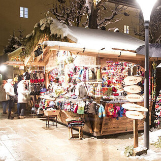 A festive Christmas market with a wooden stall offering colorful handcrafted items. The surroundings are covered in snow and adorned with decorative lights. | © Sternadvent Salzburg