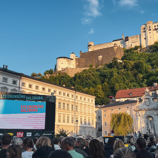 A crowd is sitting in front of a large screen displaying a movie or sports event. In the background, an impressive castle stands on a green hill. | © Tourismus Salzburg GmbH / B. Brunauer