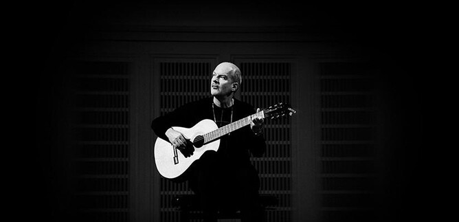 A musician is sitting on a chair and playing guitar. The scene is in black and white and radiates a calm atmosphere. | © Laura Pleiffer