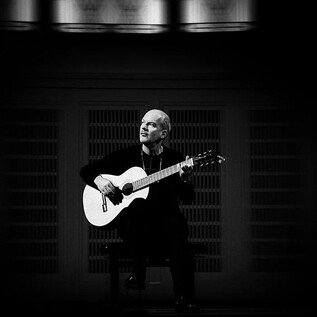A musician is sitting on a chair and playing guitar. The scene is in black and white and radiates a calm atmosphere. | © Laura Pleiffer