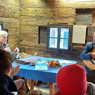 A cozy scene in a wooden cabin. A man is playing guitar while women listen with enthusiasm. | © Salzburger Freilichtmuseum