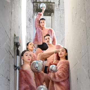 A group of people in pink furs stands in a narrow, gray corridor. They are holding silver objects in their hands and posing for the photo. | © SLT / Christian Krautzberger