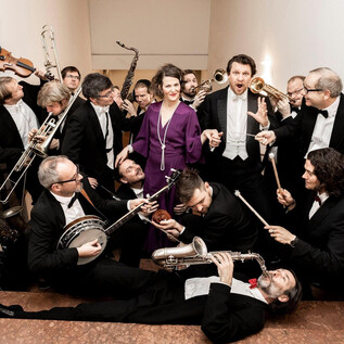 A large group of musicians in elegant evening attire plays various instruments. A woman in a purple dress stands at the center of the scene. | © Erika Mayer
