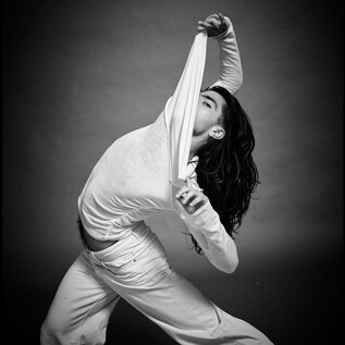 A person in a white top is stretching in a dynamic pose. The background is dark, which puts the focus on the movement and the expression. | © Chris Rogl