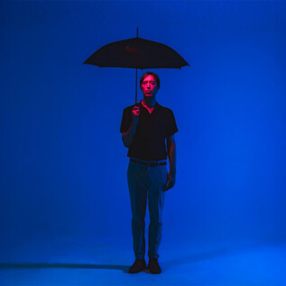 A person stands with an umbrella in front of a blue illuminated background. They are wearing a black top and have a neutral posture. | © Neil Hoare