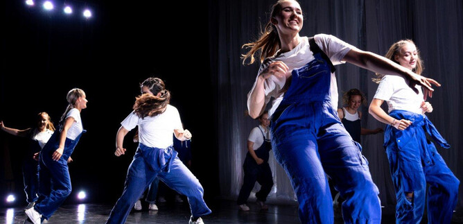 A group of dancers in blue overalls performs an energetic choreography on a stage. The atmosphere is vibrant with colorful lights in the background. | © Cornelia Pirchner