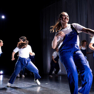 A group of dancers in blue overalls performs an energetic choreography on a stage. The atmosphere is vibrant with colorful lights in the background. | © Cornelia Pirchner