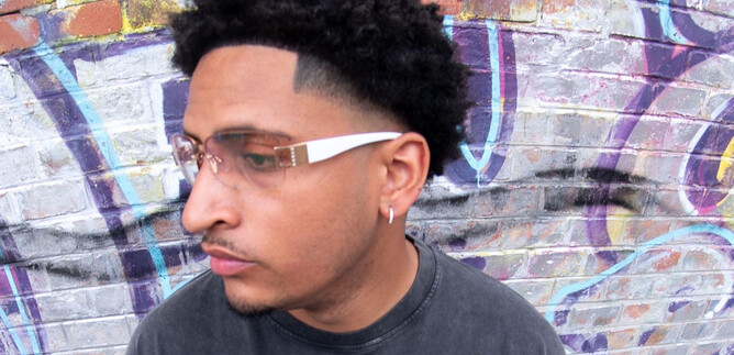 A young man with curly hair and fashionable glasses stands in front of a colorful graffiti wall. He looks thoughtfully to the side. | © Timmi Adesanya