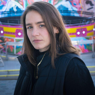 A young woman stands in an amusement park with a colorful background. She has short brown hair and is wearing a black jacket. | © Jennifer Fasching