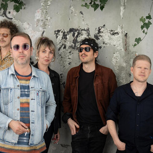 Five musicians stand in front of a gray wall with graffiti. They are smiling and look relaxed in their fashionable clothing. | © Magdalena Fuchs