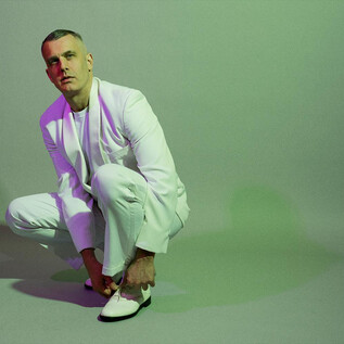 A man in a white outfit is sitting on the floor putting on his shoes. The background is in soft shades of green and purple. | © Max Zerrahh
