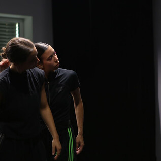 Two dancers in dark clothing stand in a room and seem to have a close interaction. The background is simple and creates a focused atmosphere. | © Dušana Baltic