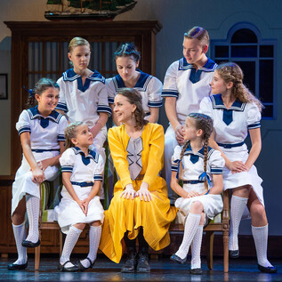 A cheerful scene from a play with children in white uniforms. A woman in a yellow dress sits in the middle and smiles. | © Anna Maria Löffelberger