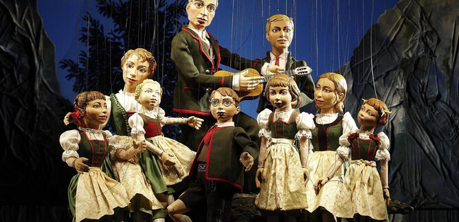 A group of dolls in traditional costumes stands on a stage. One doll is playing the guitar while the other dolls pose around her. | © Salzburger Marionettentheater