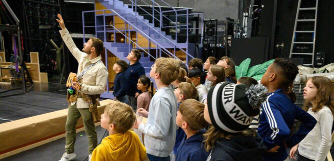 A group of children is watching with interest a teacher who is pointing at a stage. In the background, stairs and props can be seen. | © SLT_Raffael Holzinger