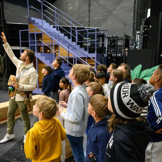 A group of children is watching with interest a teacher who is pointing at a stage. In the background, stairs and props can be seen. | © SLT_Raffael Holzinger