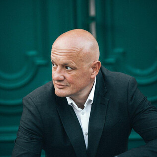 A bald man is wearing a black suit and looking thoughtfully. In the background, green doors are visible. | © Maria Tiefenbacher-Schöndorfer
