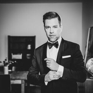 A well-dressed man in a suit with a bow tie stands in a room. The background features furniture and a globe decoration. | © Moritz Künster