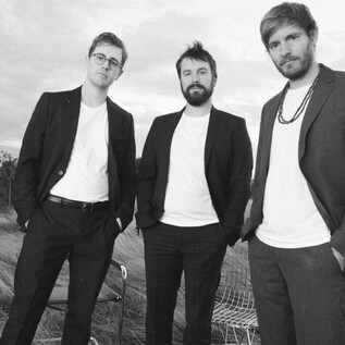 Three men in suits stand in an open field. They appear self-confident and are photographed in a modern, artistic black-and-white style. | © Julian Hagen