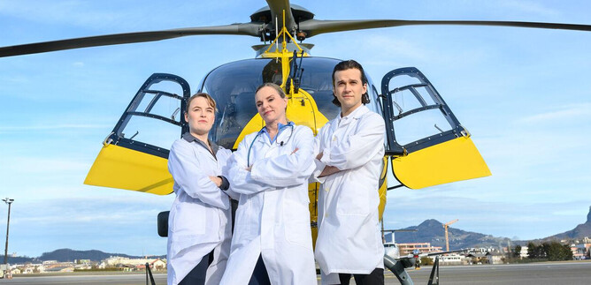 Three people in white coats are standing in front of a helicopter. The sky is clear and the surroundings are an urban landscape. | © SLT / Tobias Witzgall