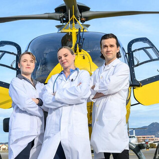 Three people in white coats are standing in front of a helicopter. The sky is clear and the surroundings are an urban landscape. | © SLT / Tobias Witzgall