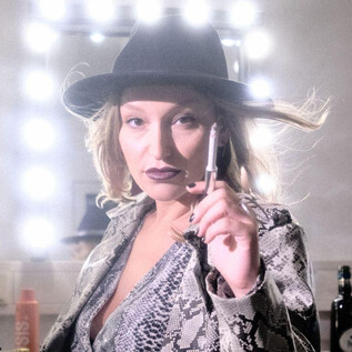 A woman with a black hat and a snake-patterned jacket holds a pen in her hand. The background is illuminated with mirror lights. | © Christopher Glanzl