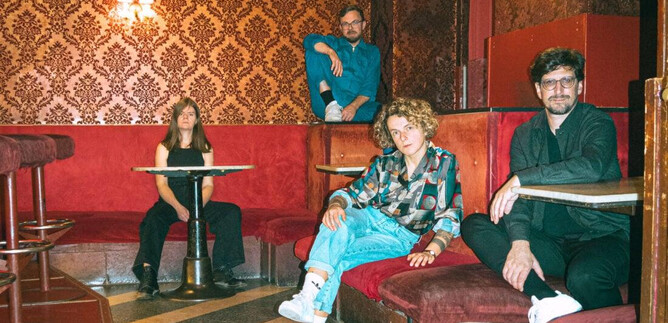 A group of four people is sitting and standing in a stylishly decorated room with red decor. The atmosphere is relaxed and creative. | © Julia Freytag