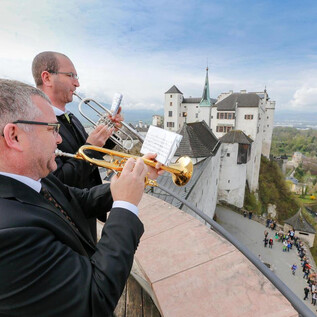 Two musicians are playing trumpets with an impressive castle in the background. The scene showcases a blend of culture and history against a picturesque landscape. | © www.salzburg-burgen.at