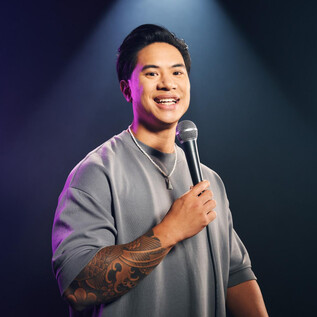 A young man with a microphone smiles at the camera. He is wearing a gray sweatshirt and is standing in front of a dark, illuminated background. | © Marvin Ruppert