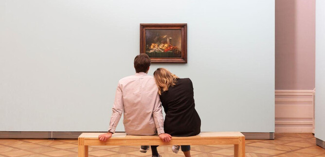 A couple sits on a bench in an art museum, gazing at a painting. The room is designed in a minimalist style and features a soft color palette. | © DomQuartier