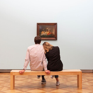 A couple sits on a bench in an art museum, gazing at a painting. The room is designed in a minimalist style and features a soft color palette. | © DomQuartier